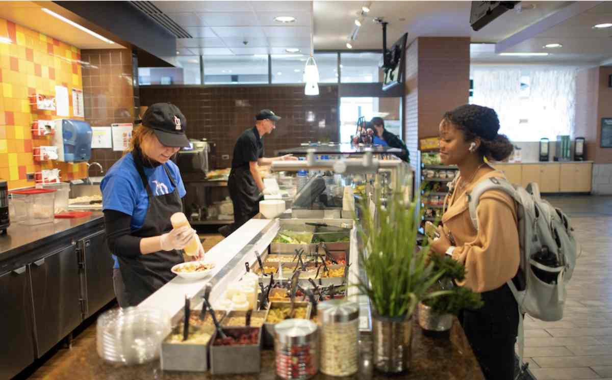 A girl smiling as she orders a salad and the worker happily making her food at a Laker Food Co. place