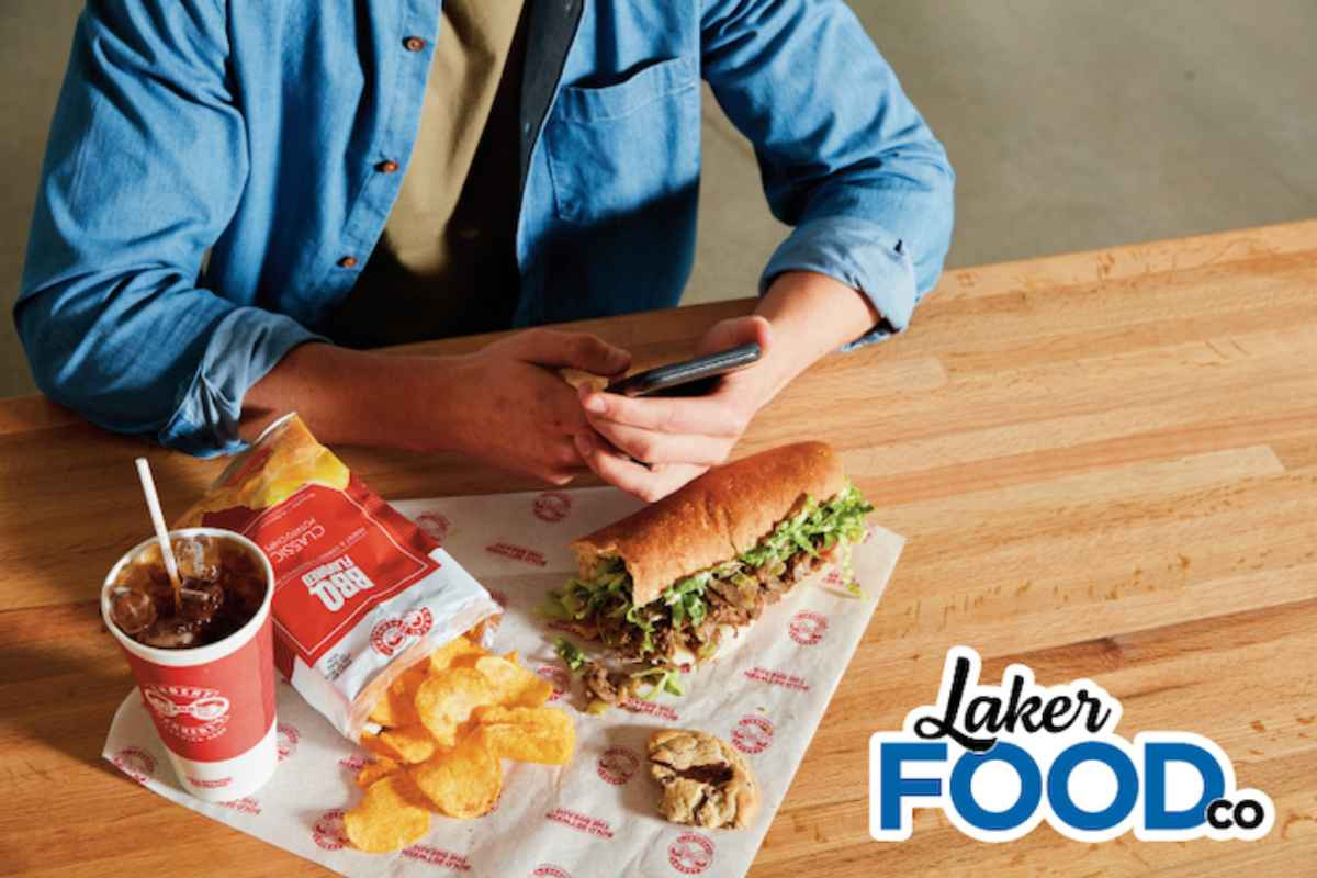 A student eating some Erbert and Gerbert's sandwhich, chips and a drink with the Laker Food Co.  logo on the bottom right