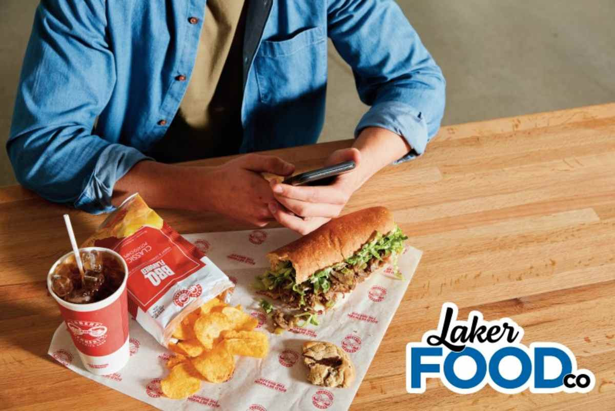 A student eating a sandwhich, drink and chips from the new restaurant on campus with the Laker Food Co. logo