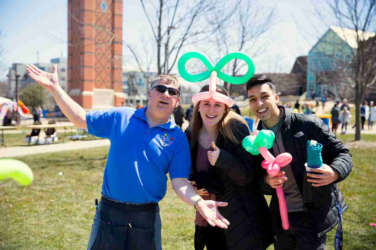 a balloon animal artist taking a photo with two students; one who has a balloon hat on