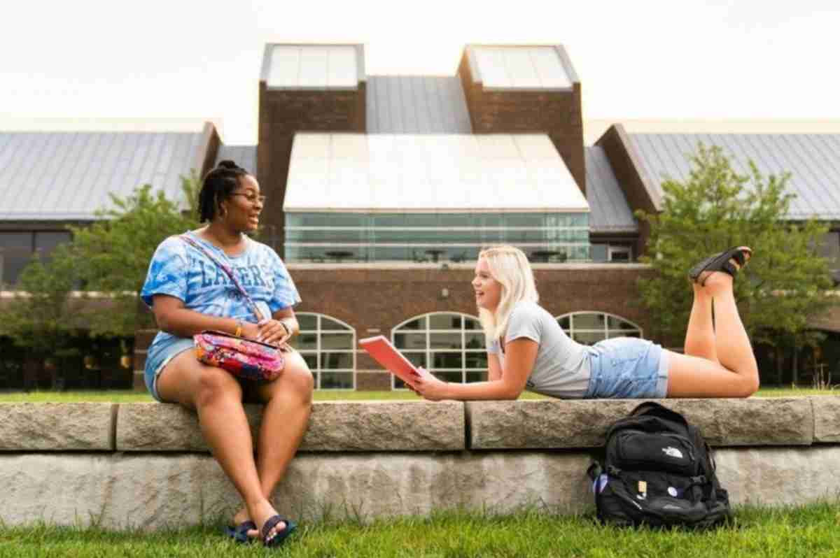 A student laying on their stomach in the grass talking to another student who is sitting