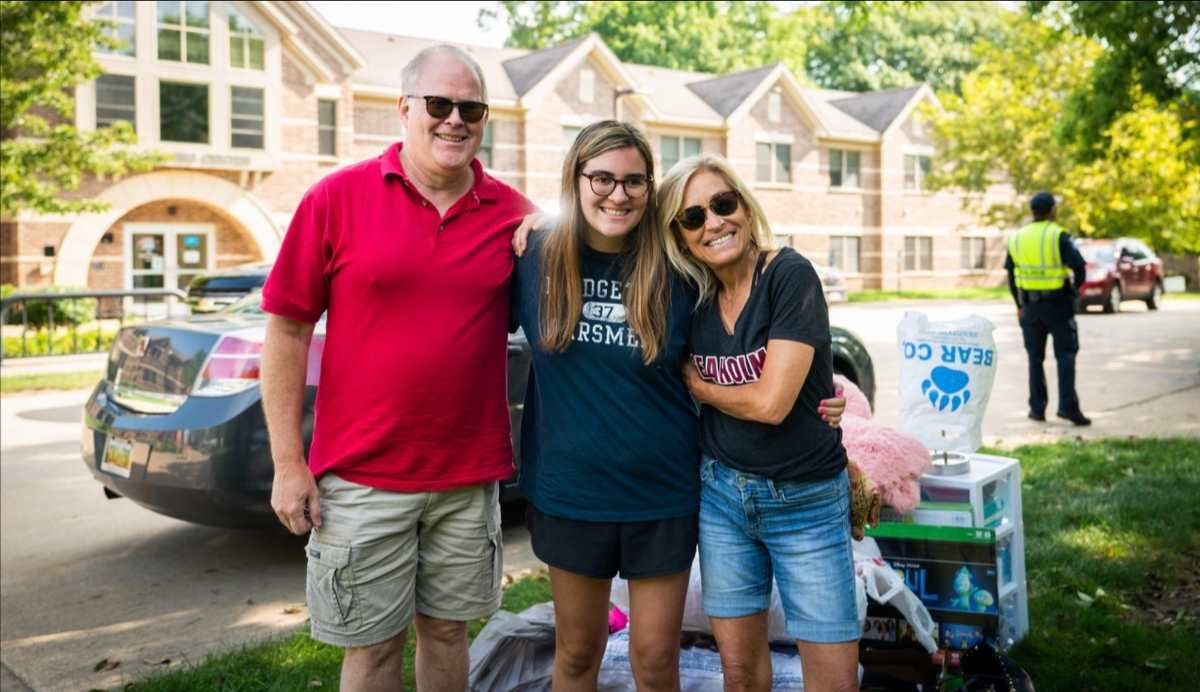 a student and their parents smiling for a parent in front of their car packed to move home.