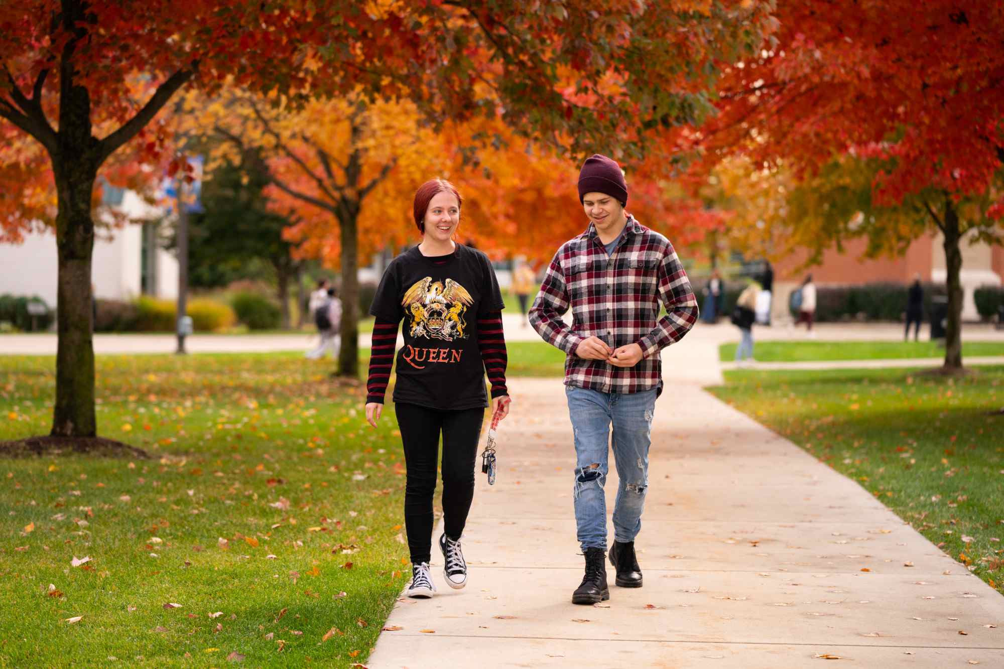 two people walking down the sidewalk with the fall leaves in the background 