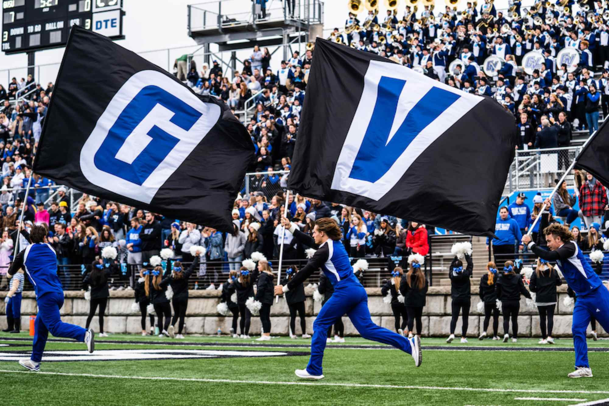 cheerleaders running down the field with flags that have the letters G and V on them during the Homecoming Football Game