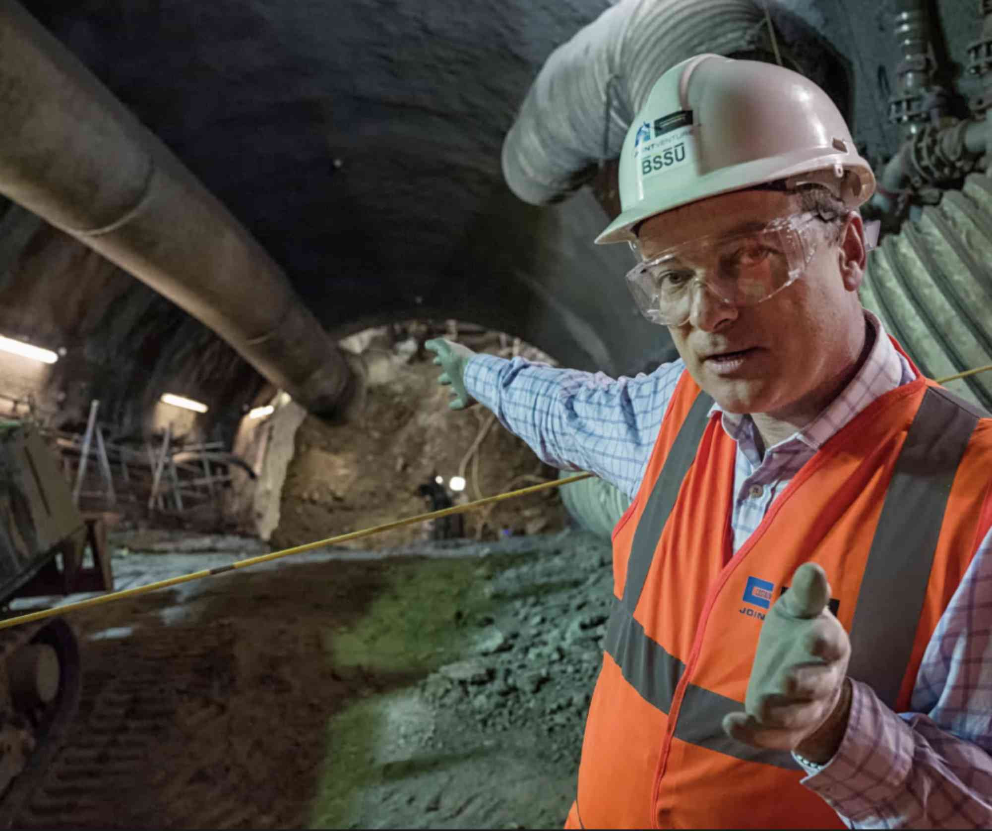 A man in an orange construction vest and a white hard had standing in an underground tunnel and pointing behind him