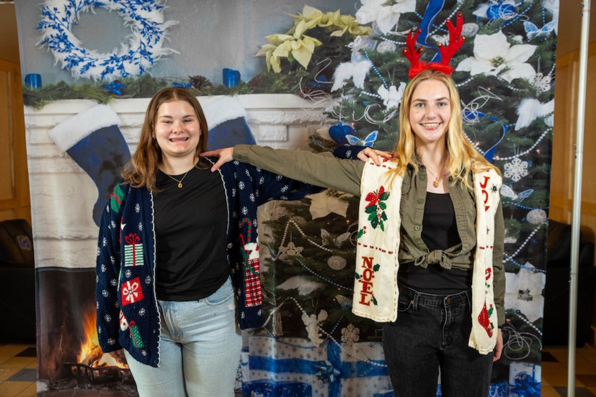 Two people awkwardly posing wearing Christmas scarves and sweaters and reindeer antlers