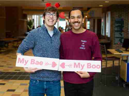 Two men smiling holding signs that say "my boo" and "my bae"