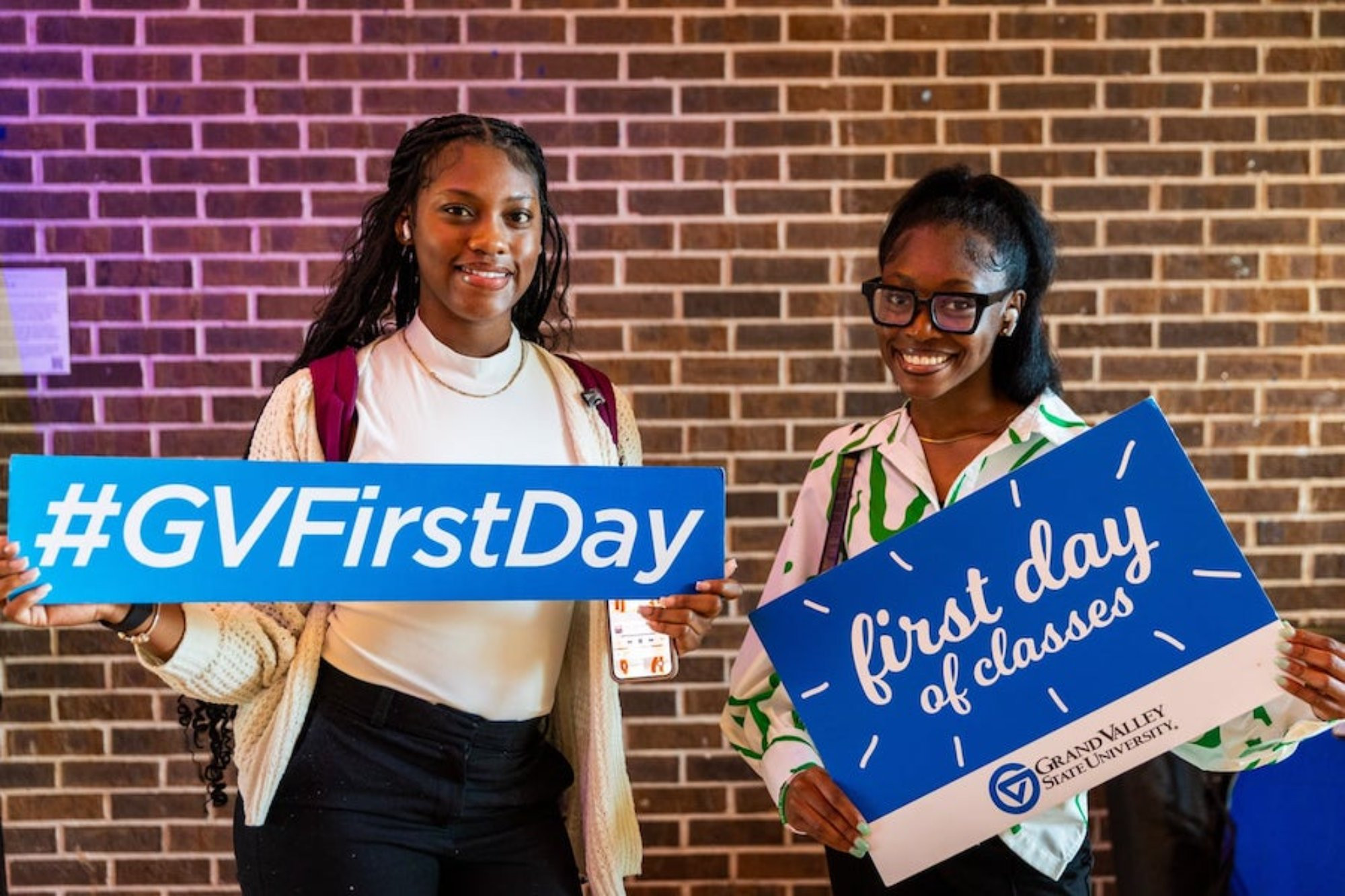 Friends posing for a first day of school photo, one girl holding a hashtag GVFirstDay sign and another holding a sign that says First Day of Classes sign with the GVSU Logo on the bottom