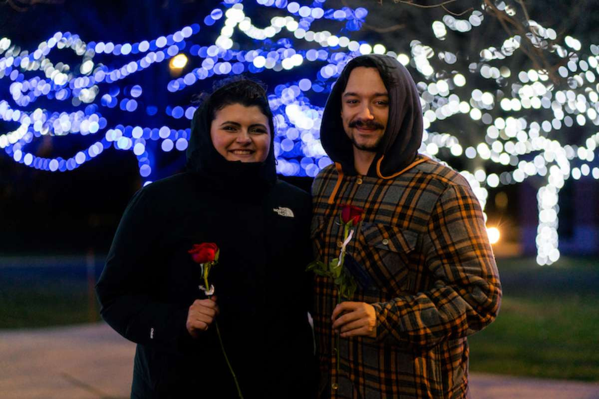 Two students wearing winter coats smiling and standing in front of trees draped with white and blue holiday lights