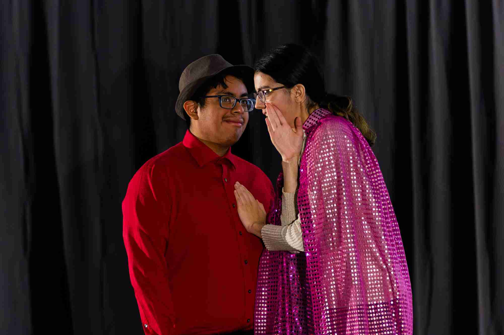 A student wearing a sparkly pink cape whispers to a student in a red shirt and black hat