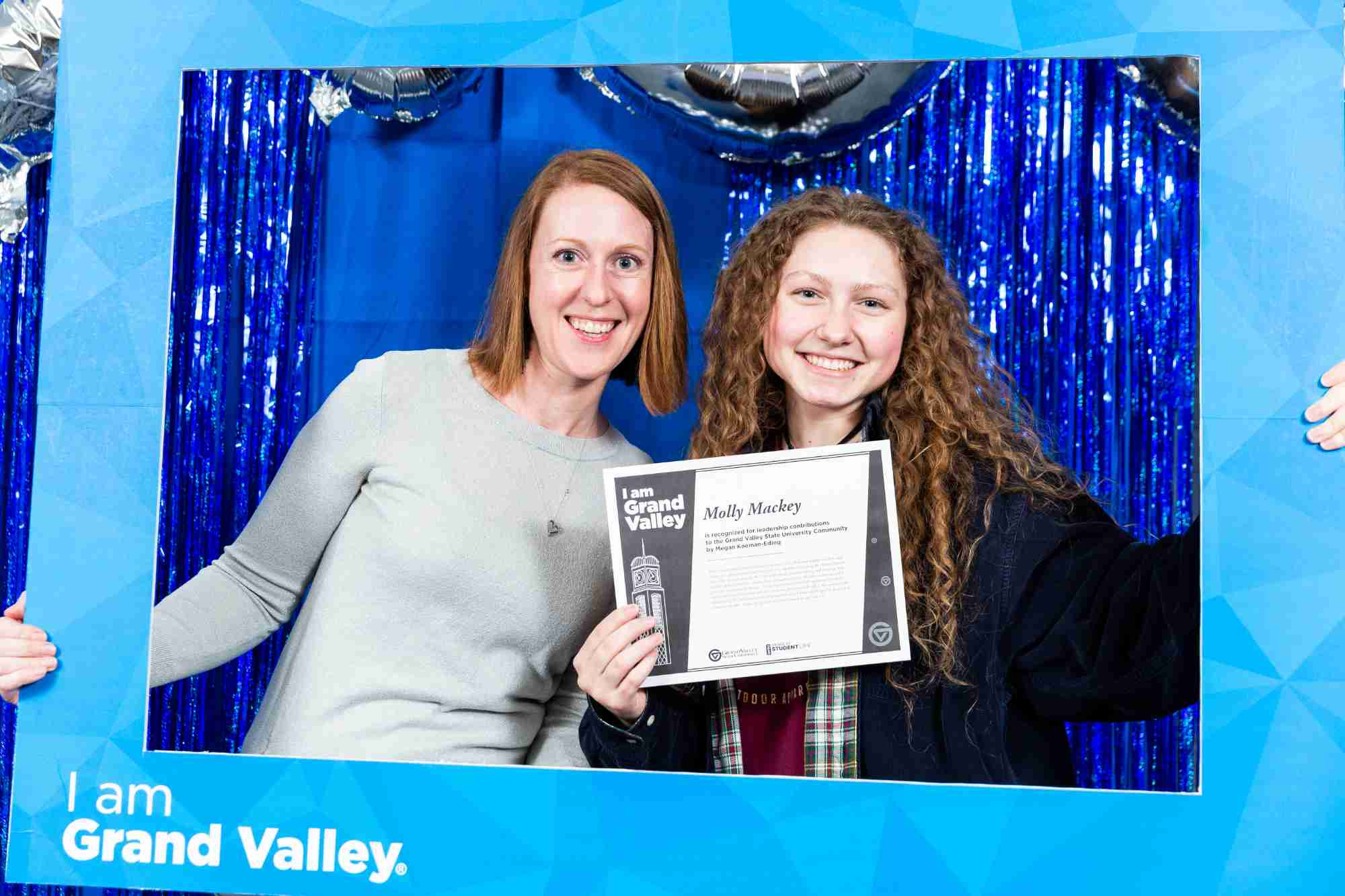 A student who received a I am Grand Valley award taking a picture with a peer behind a I Am Grand Valley photo frame prop